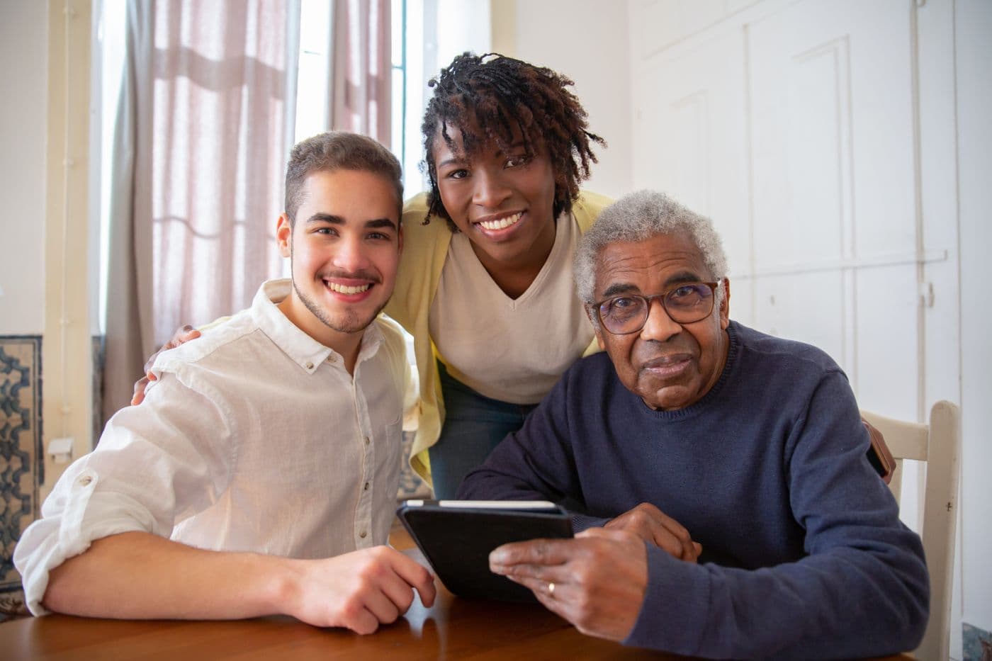 A caregiver and a young family member sitting close beside a smiling older man at a table in his home